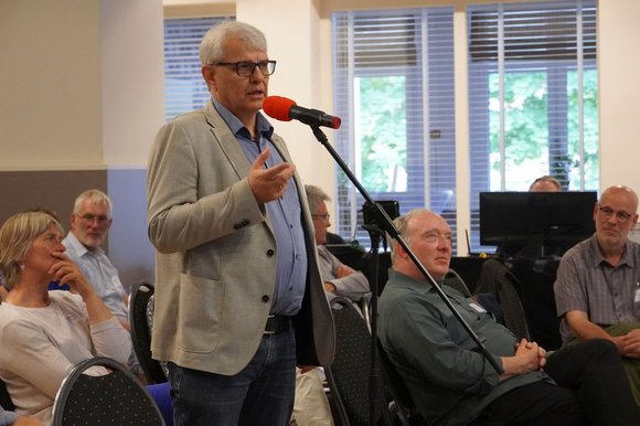 Ein Mann mit grauen Haaren steht an einem Mikrofon und spricht.