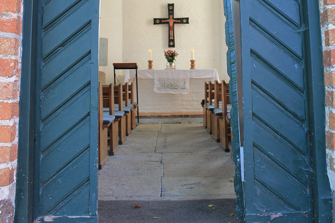 Eine offene blaue Kirchentür gibt den Blick frei zu einem Kirchenaltar auf dem zwei große, weiße, brennende Kerzen zu sehen sind. Zwischen den Kerzen steht eine runde Glasvase mit Blumen, darüber hängt ein Altarkreuz. Im vordergrund sind braune Holz-Kirchenbänke mit blauen Sitzauflagen zu sehen.