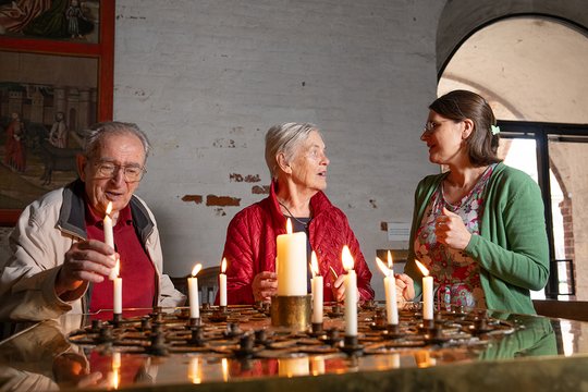  Ein älterer Herr mit Brille, rotem Pullover und heller Jacke steht links hinter einem Tisch mit brennenden Kerzen. Er steckt gerade eine weitere Kerze in die Vorrichtung. Rechts neben ihm steht eine ältere Dame mit roter Jacke, sie unterhält sich mit einer Frau, welche rechts von ihr steht. Diese trägt eine grüner Strickjacke. Sie befinden sich im Innenraum einer alten Kirche.