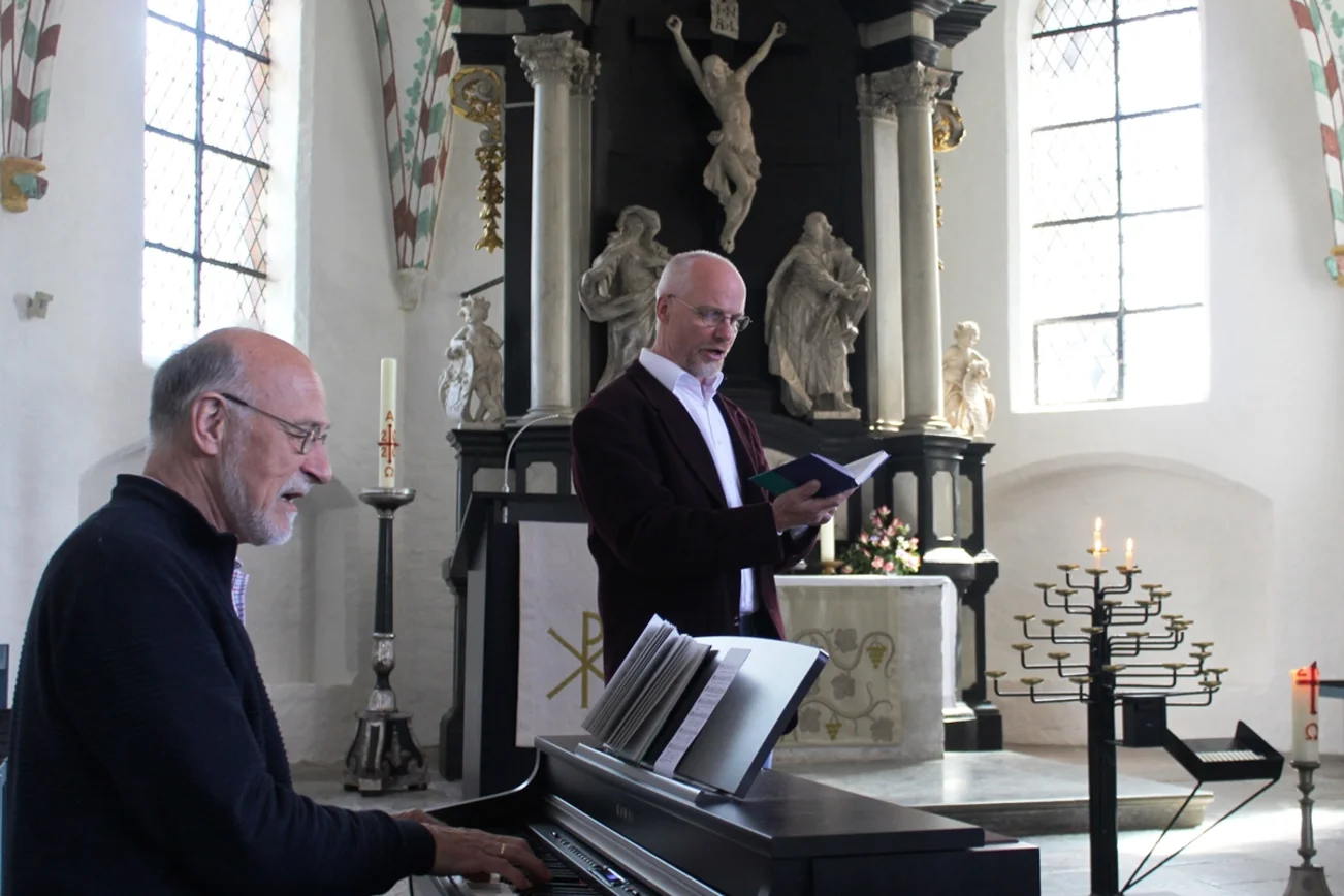Ein Mann im Profil, sitzend am Klavier. Ein Mann dahinter stehend mit Mappe in der Hand.