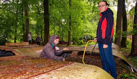 Zwei Mädchen stehen im Wald und blicken auf das Wellblechdach eines alten Anhängers. 