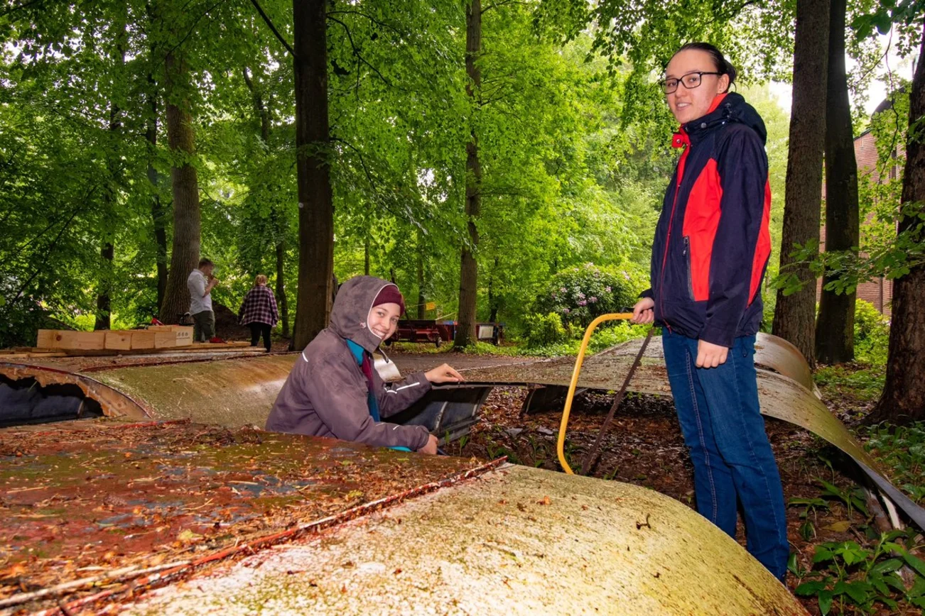 Zwei Mädchen stehen im Wald und blicken auf das Wellblechdach eines alten Anhängers. 