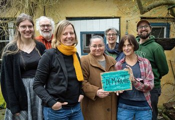 Sieben Frauen und Männer stehen als Gruppe zusammen und zeigen ein Schild mit der Aufschrift Demokratie. - Copyright: Gesa Hollaender