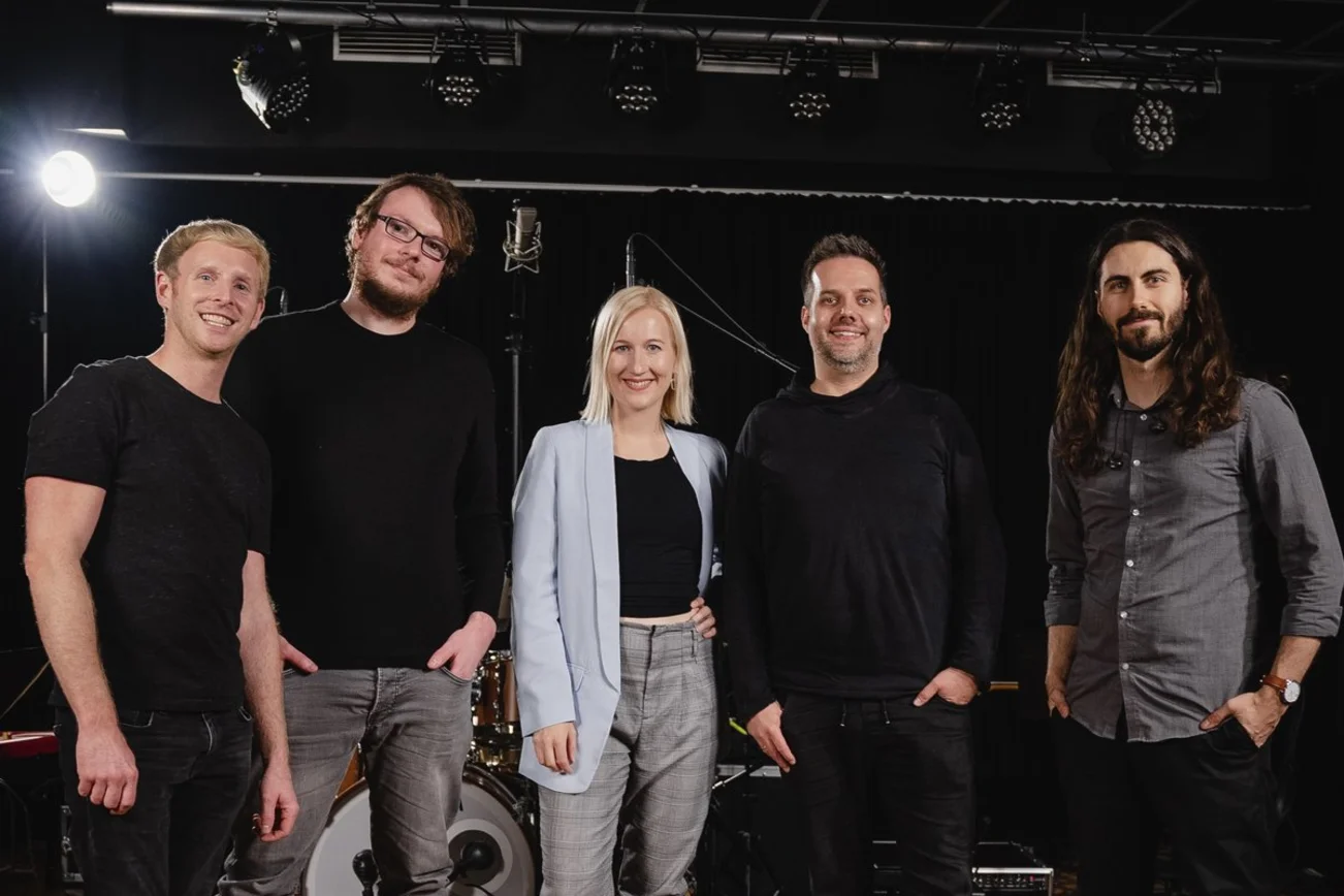 Eine Gruppe von Menschen steht in einem Probenraum und lächelt in die Kamera. 