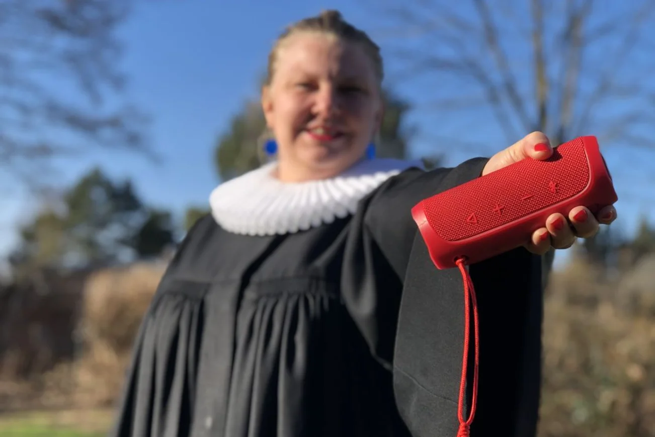 Eine Frau im Talar mit weißem Kragen hält eine rote Bluetooth-Box in die Kamera.