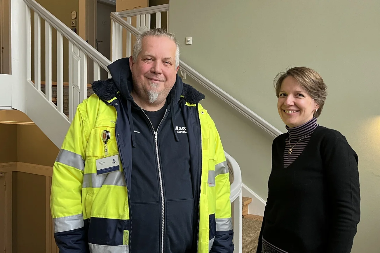 Ein Mann mit gelber Warnjacke steht neben einer Frau mit schwarzem Pullover vor einem weißen Treppengeländer.