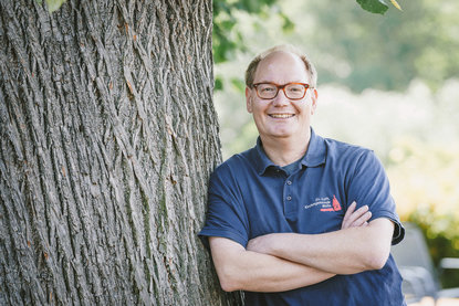 Mann mit Brille lehnt an einem breiten Baum und guckt freundlich in die Kamera. - Copyright: Guido Kollmeier