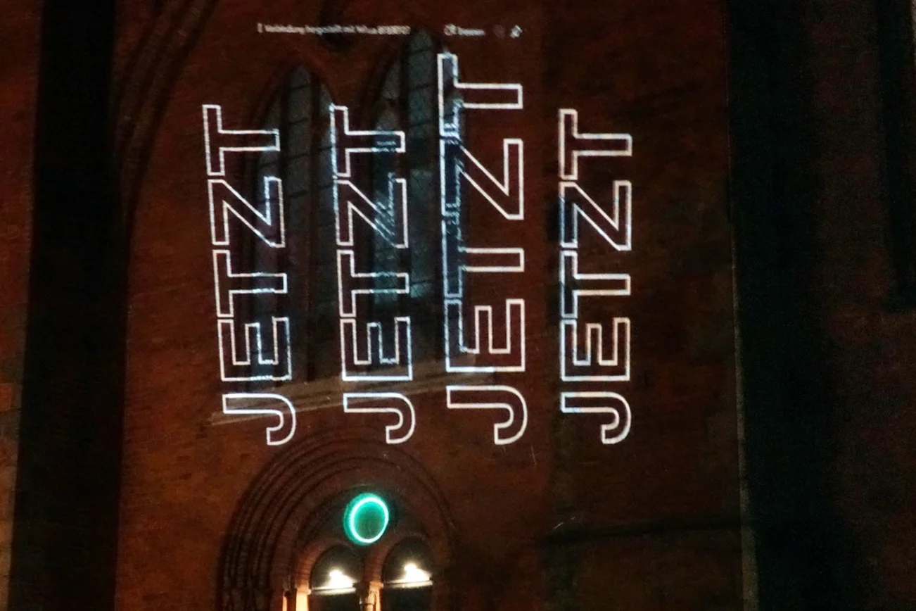 Das Wort Jetzt wird in auf die Backsteinfassade einer Kirche projeziert.