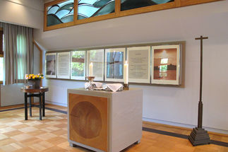 Andachtsraum der Universitätsklinik Schleswig-Holstein - Der Altar ist mir Kerzen und zugedecktem Abendmahl gedeckt. An der Wand hängen verschiedene Bilder und Texte. Auf der linken Seite steht ein kleiner runder Tisch auf dem eine Vase mit verschieden farbenen Rosenbunter Rosen steht. Rechts vom Altar und den Bildern steh ein schlanken hohes Kreuz.