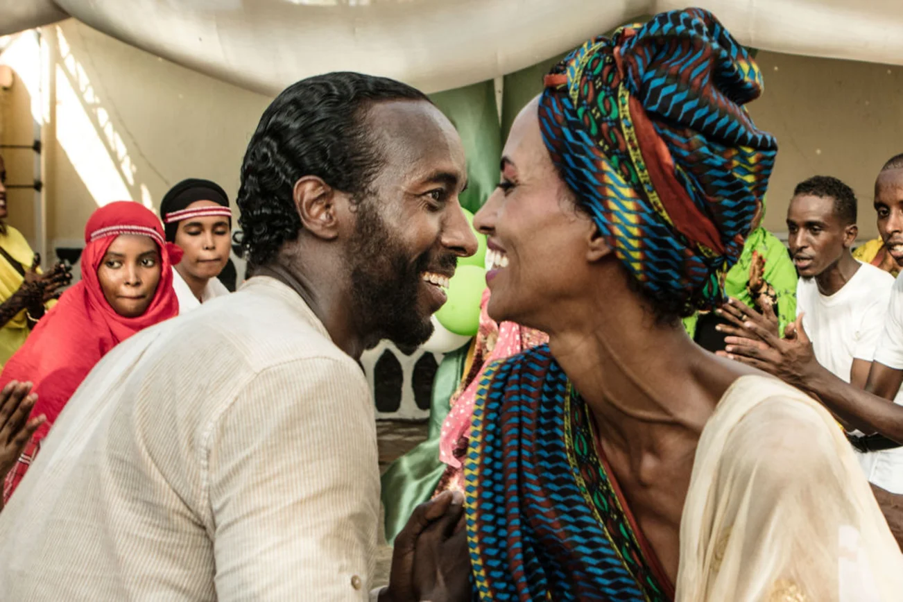 Ein Mann und eine Frau sitzen sich lächelnd gegenüber, ringsum sind weitere fröhliche Menschen zu sehen. 