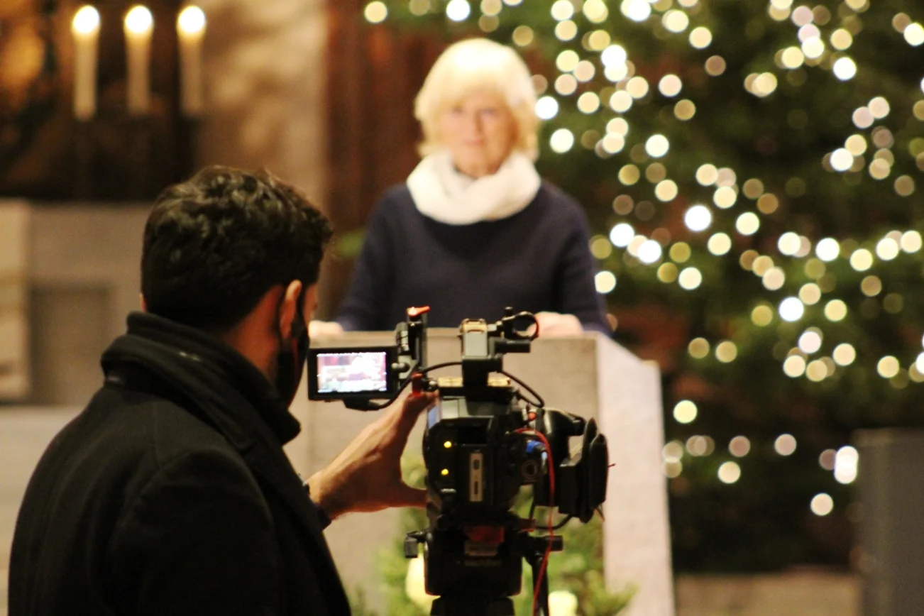 Im Vordergrund eine Kamera. Sie ist auf eine Frau an einem Rednerpult gerichtet. Im Hintergund ein Kirchraum mit Tannenbaum