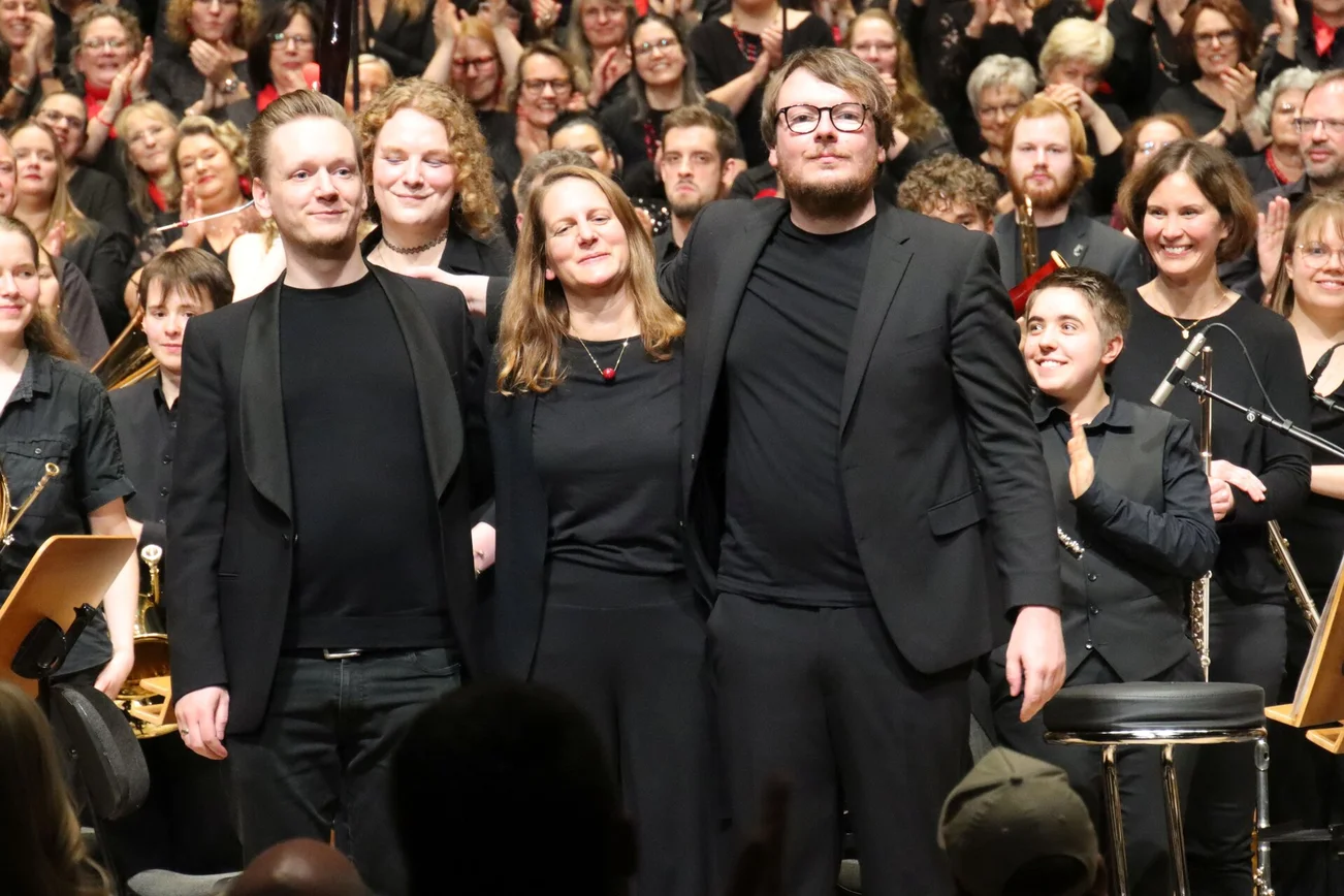 Zwei Männer und eine Frau in schwarzer Kleidung stehen vor einem Chor auf der Bühne
