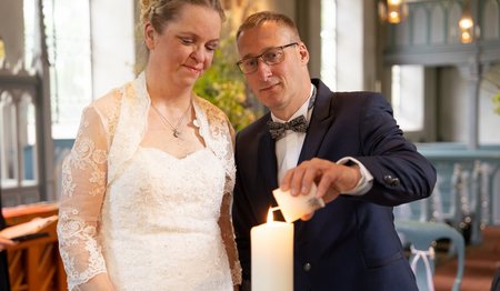 Ein Brautpaar steht vor einer Kerze. Der Mann entzündet ein Licht an der Kerze.