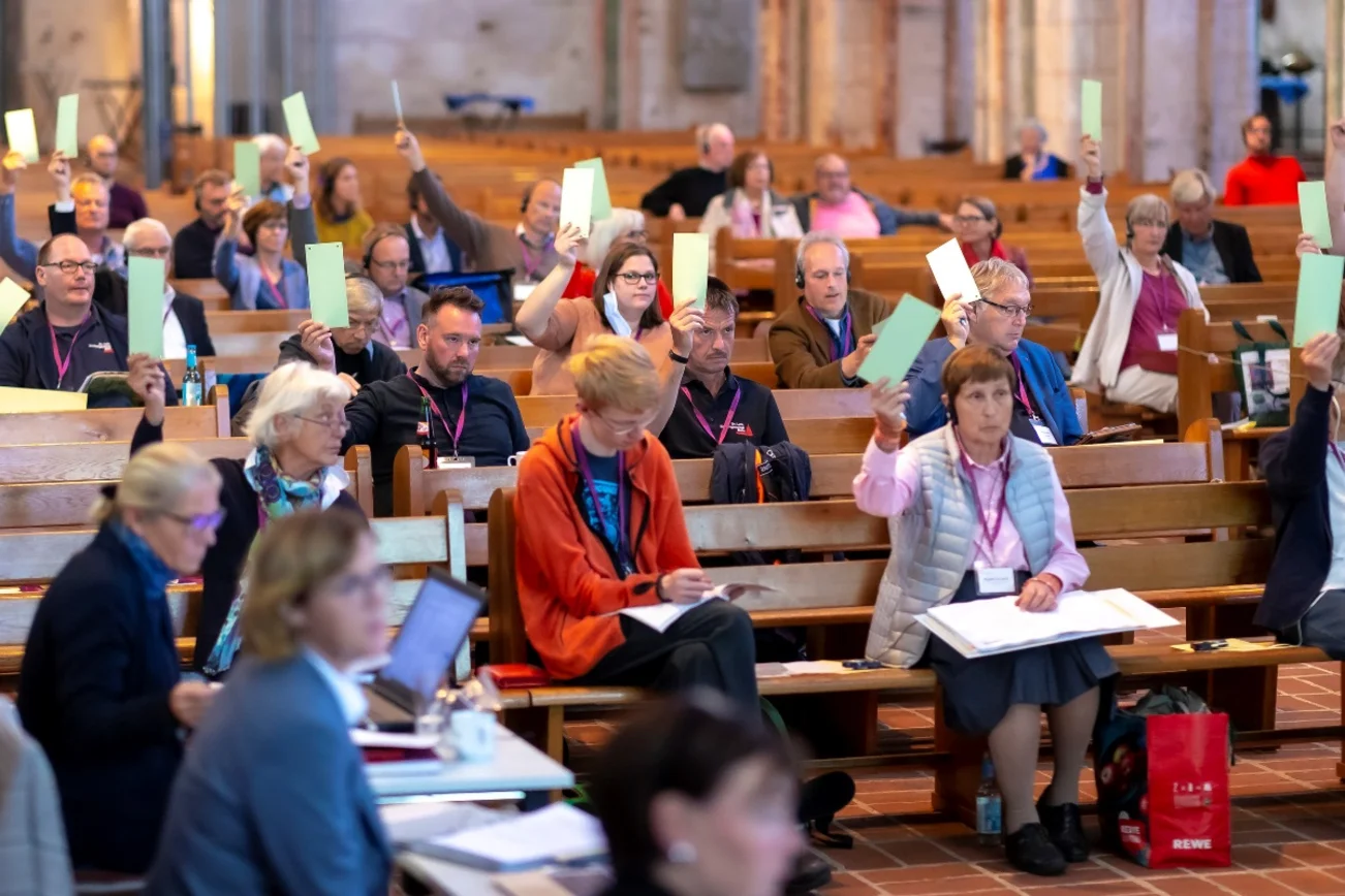 Menschen sitzen auf Abstand auf Kirchenbänken und heben grüne Karten nach oben.