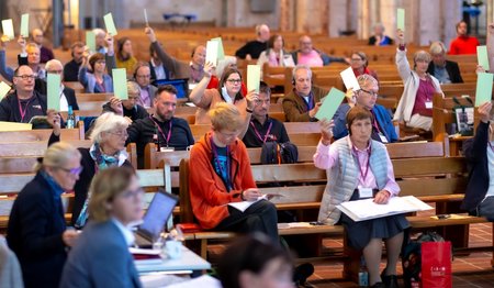 Menschen sitzen auf Abstand auf Kirchenbänken und heben grüne Karten nach oben.