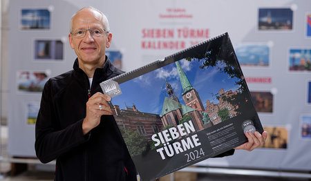 Fotograf Rainer Jensen hält den Sieben Türme Kalender 2024 in den Händen, im Hintergrund die Ausstellungstafel zur Verlosung der Sonderedition 2025 im Dom