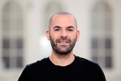 Ein Mann steht vor einer hellen, weißen Wand mit 3 hohen Fenstern, welche nur unscharf zu erkennen sind. Der Mann hat einen 3 Tage-Bart und ganz kurze Haare und lächelt in die Kamera. Er trägt ein schwarzes Shirt. - Copyright: Anne Oschatz