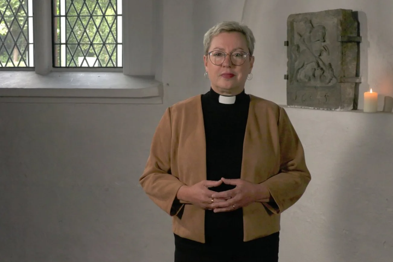 Eine Frau steht in einer Kirche und blickt ernst in die Kamera. 