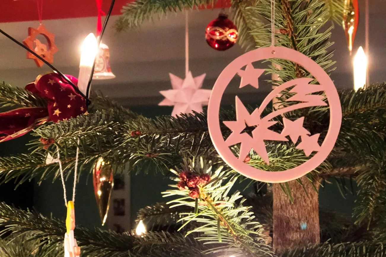Ein Christbaum mit Schmuck, im Vordergrund ein Holzanhänger mit Sternschnuppe.