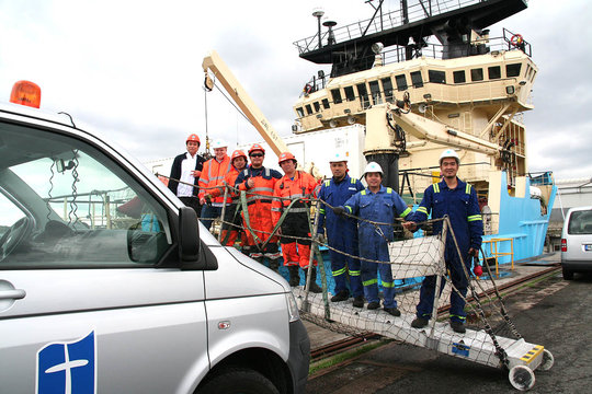 Die Crew eines Schiffes steht auf dem mobilen Landungssteg zwischen Schiff und Festland und schaut in die Kamera. Links im Bild, direkt vor dem Schiff, sieht man das Führerhaus vom Auto der Seemannsmission.