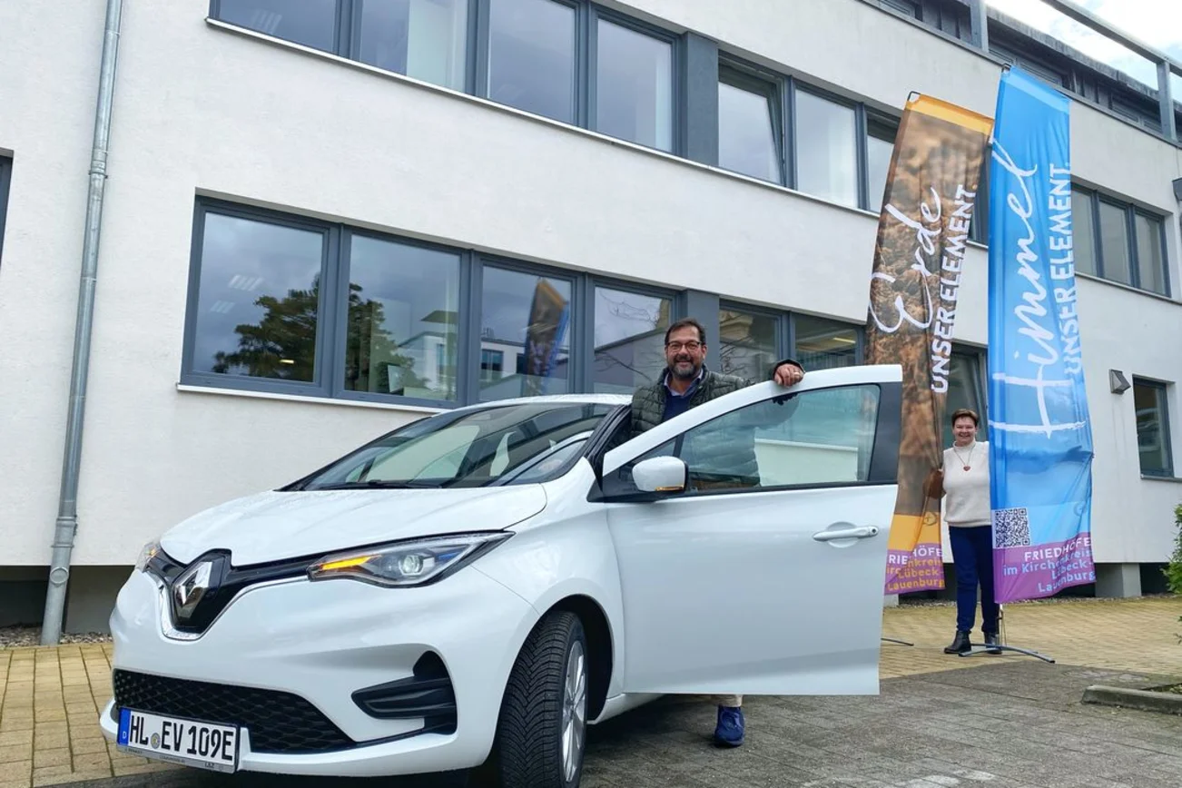 Zwei Personen stehen vor einem weißen E-Auto und lächeln in die Kamera. 
