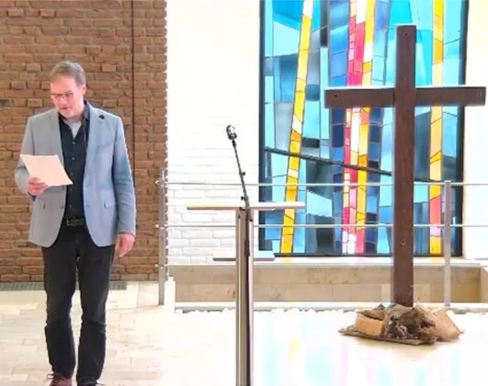 Ein Mann hält einen Zettel in der Hand auf den er schaut, rechts neben ist steht ein Mikrophon und ein Stehpult, weiter rechts ein großes Holzkreuz und ganz im Hintergrund ist ein buntes Kirchenfenster