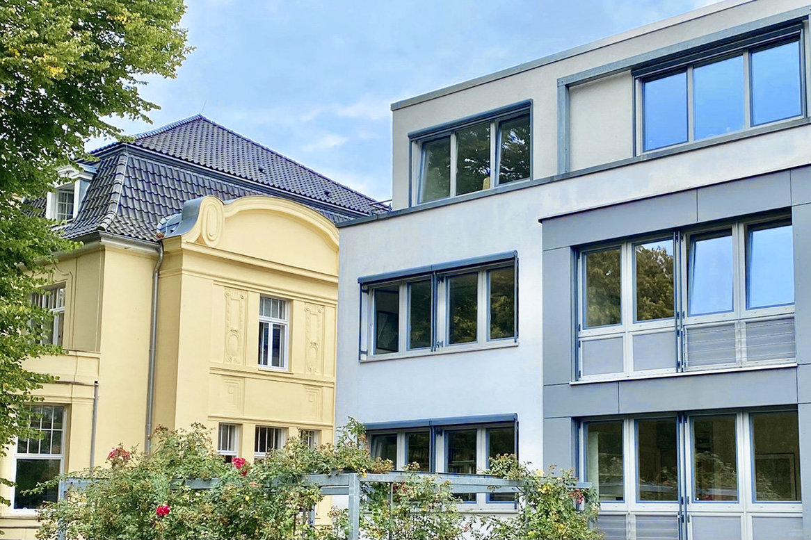 Alt- und Neubau der Kirchenkanzlei - Auf der linken Seite der Altbau mit gelber Fassade und dunklem Dach, rechts der Neubau in Grautönen und Flachdach