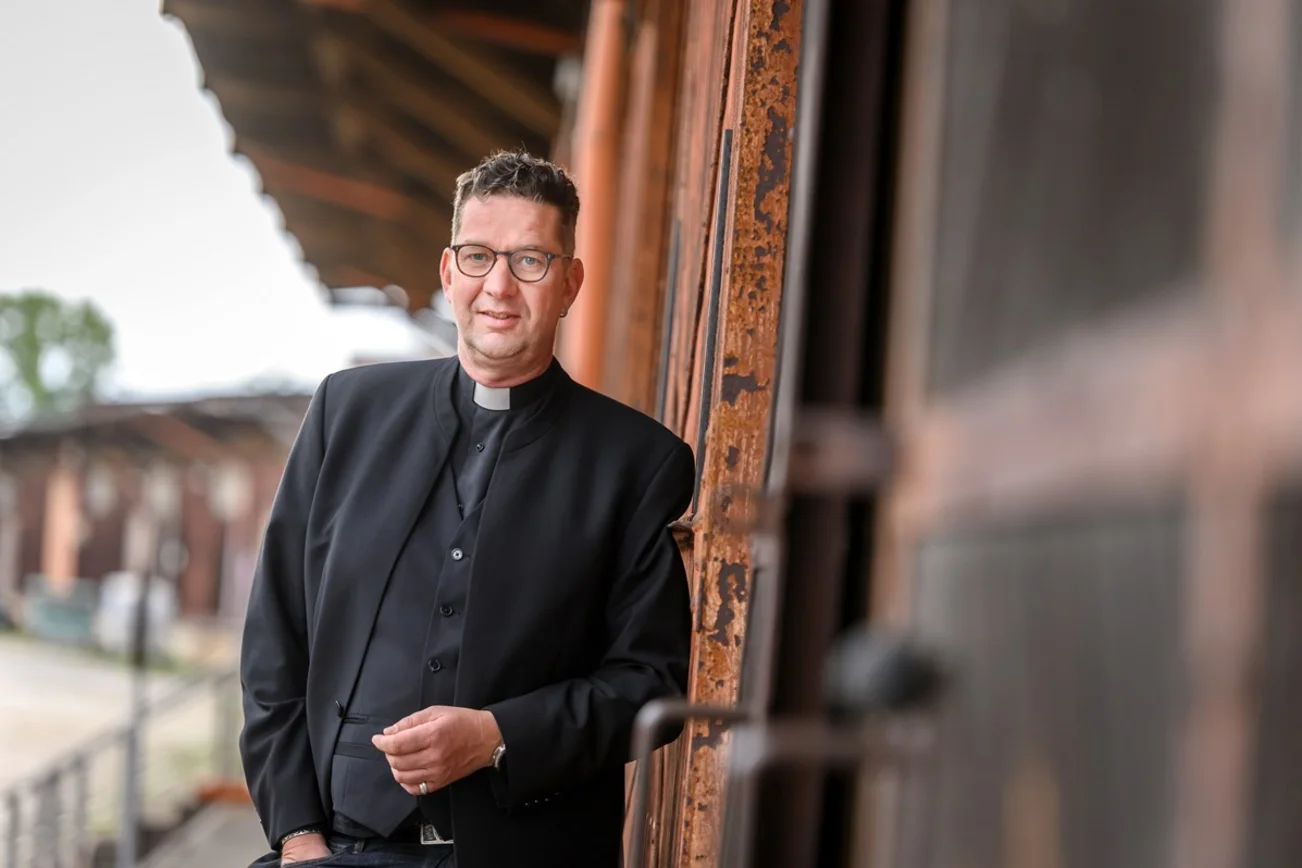 Ein Mann im schwarzen Anzug, mit Brille lehnt an der roten Holzwand einer Remise. 
