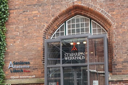 Eingangsbereich aus Glas an einer roten Backsteinfassade mit der Aufschrift St. Marienwerkhaus und links ein Schild Seniorenakademie Lübeck