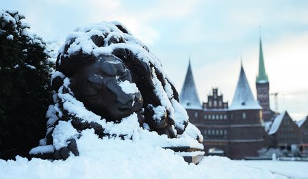 Der Steinlöwe vor dem Holstentor Lübeck ist in einer schlafenden Position. Er ist mit Schnee bedeckt, ebenso das Holstentor im Hintergrund und rechts daneben St. Petri.