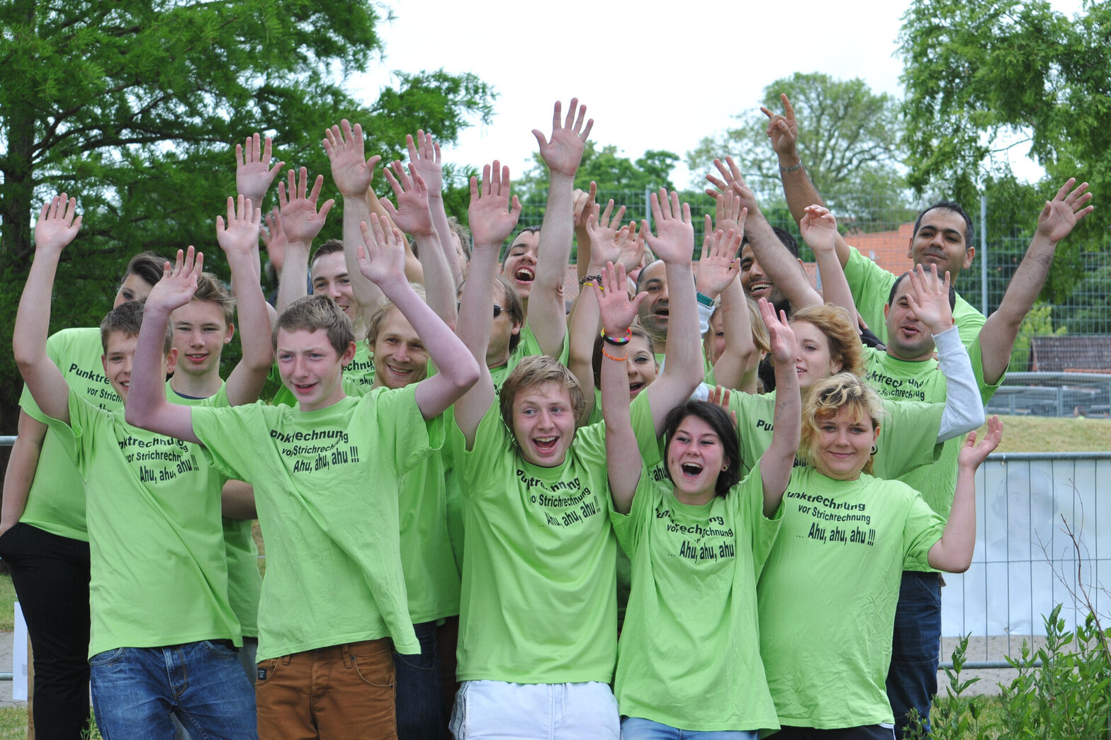 Mehrere junge Leute mit grünen T-Shirts stehen in mehreren Reichen dicht hintereinander. Sie halten die Arme hoch und schauen fröhlich in die Kamera