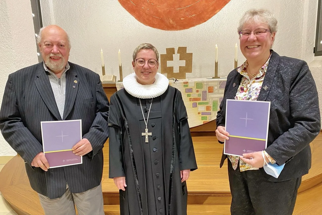 Drei Personen stehen lächelnd vor einem Altar. 