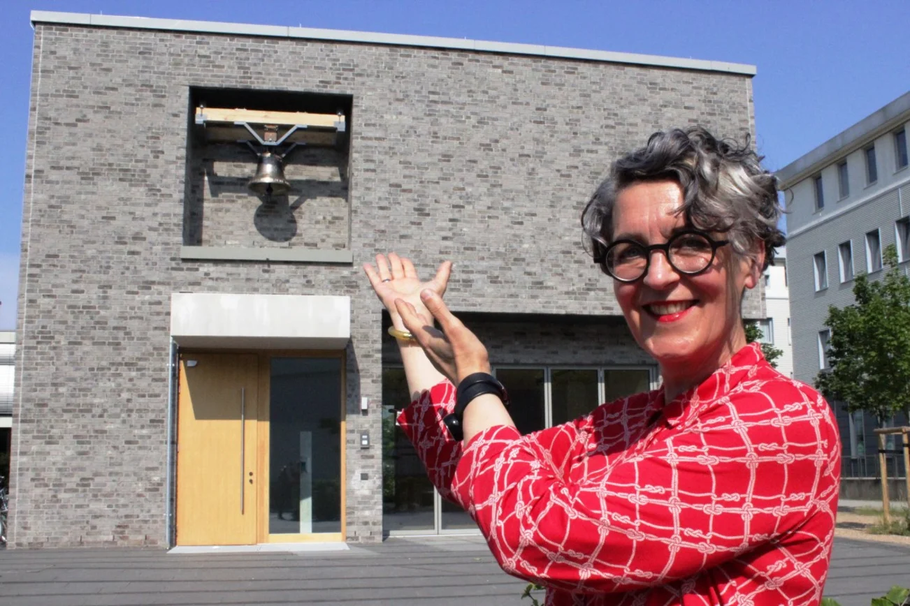 Eine Frau zeigt auf ein Haus. Blauer Himmel, am rechten Bildrand ein Baum.