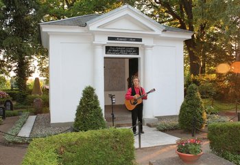 Ein Mann steht mit seiner Gitarre vor einem Grabmal. - Copyright: Hans W. Kuhlmann