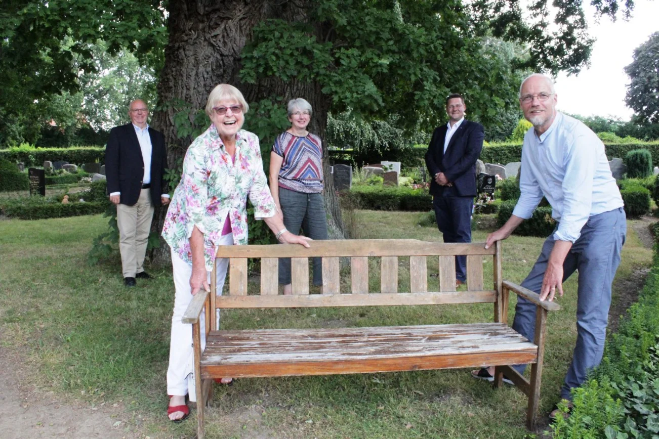 Funf Männer und Frauen auf Abstand rund um eine Holzbank. Im Hintergrund ein Baum.