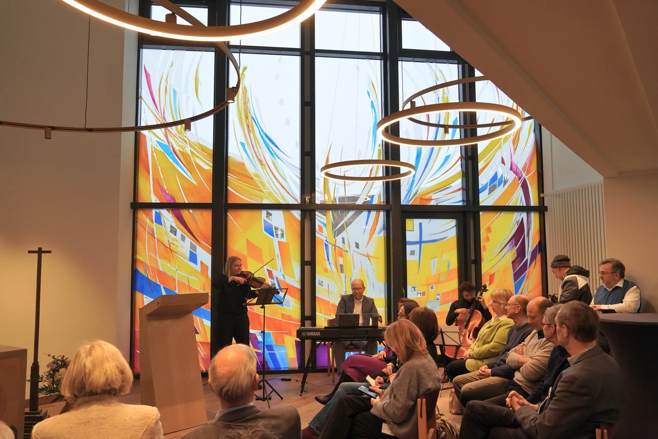 Ein Raum mit großen bunten Fenstern, vielen sitzenden Menschen und drei musizierendenMusikern.
