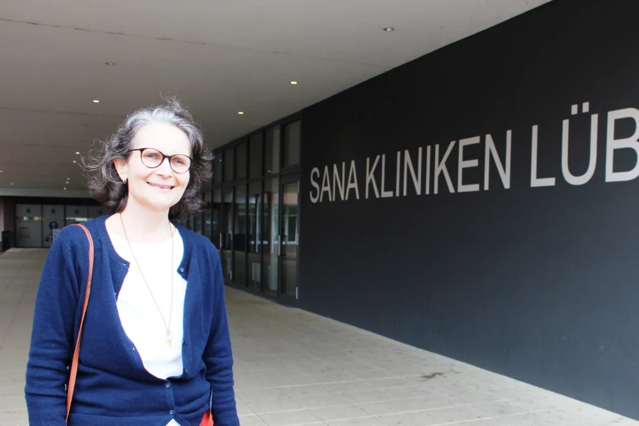 Eine Frau mit Brille, weißer Bluse und blauer Jacke steht vor einem Eingangsbereich. Im Hintergrund ist die Aufschrift Sana Kliniken Lübeck zu lesen.