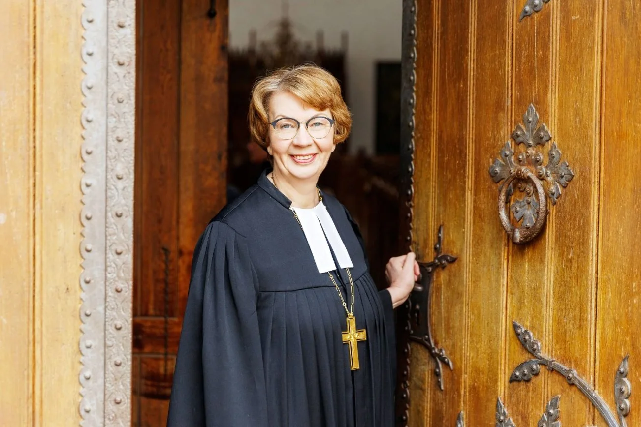 Eine Frau im Talar mit Bischofskreuz kommt aus einer Kirchentür heraus. Sie lächelt freundlich.
