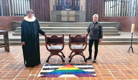 Zwei Personen stehen in einer Kirche neben zwei freien Stühlen. 