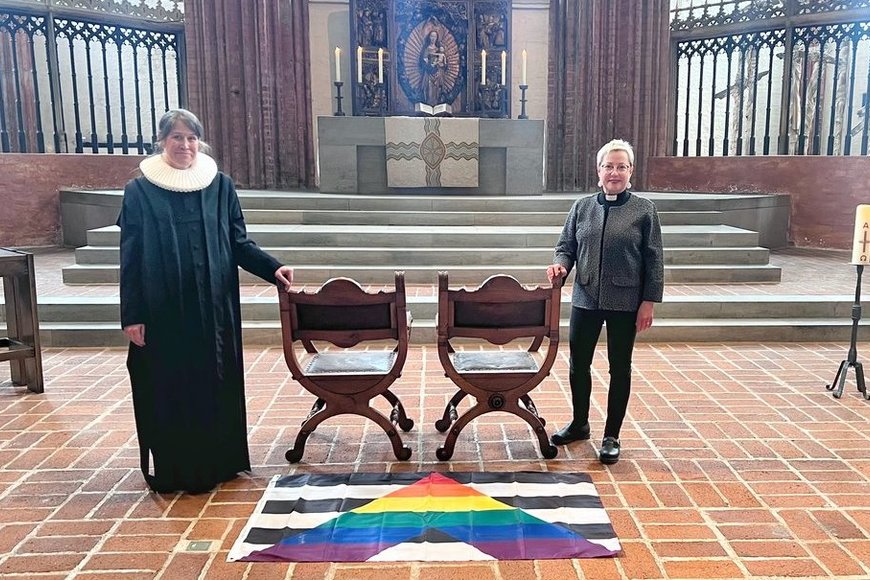 Zwei Personen stehen in einer Kirche neben zwei freien Stühlen.  - Copyright: KKLL