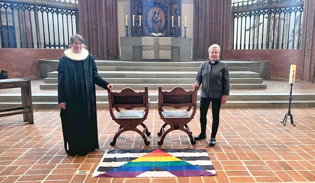 Zwei Personen stehen in einer Kirche neben zwei freien Stühlen. 