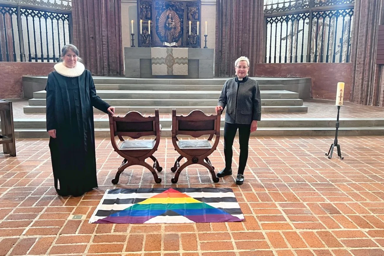 Zwei Personen stehen in einer Kirche neben zwei freien Stühlen. 
