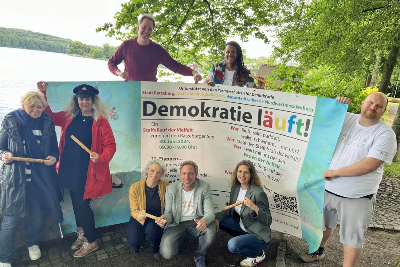 Eine Gruppe Menschen steht mit einem großen Plakat an einem See.