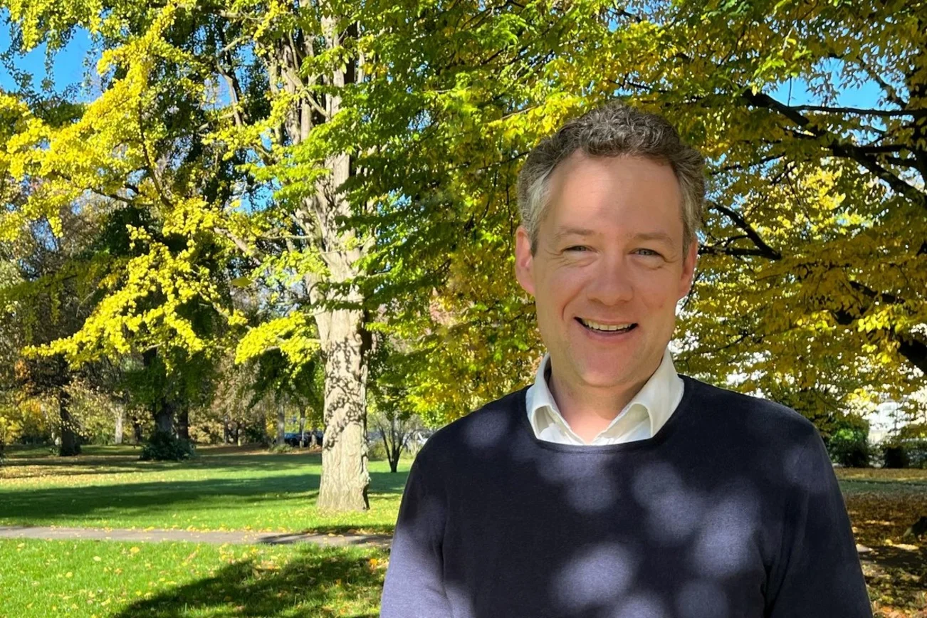 Mann steht vor einem Laubbaum und lächelt  in die Kamera.