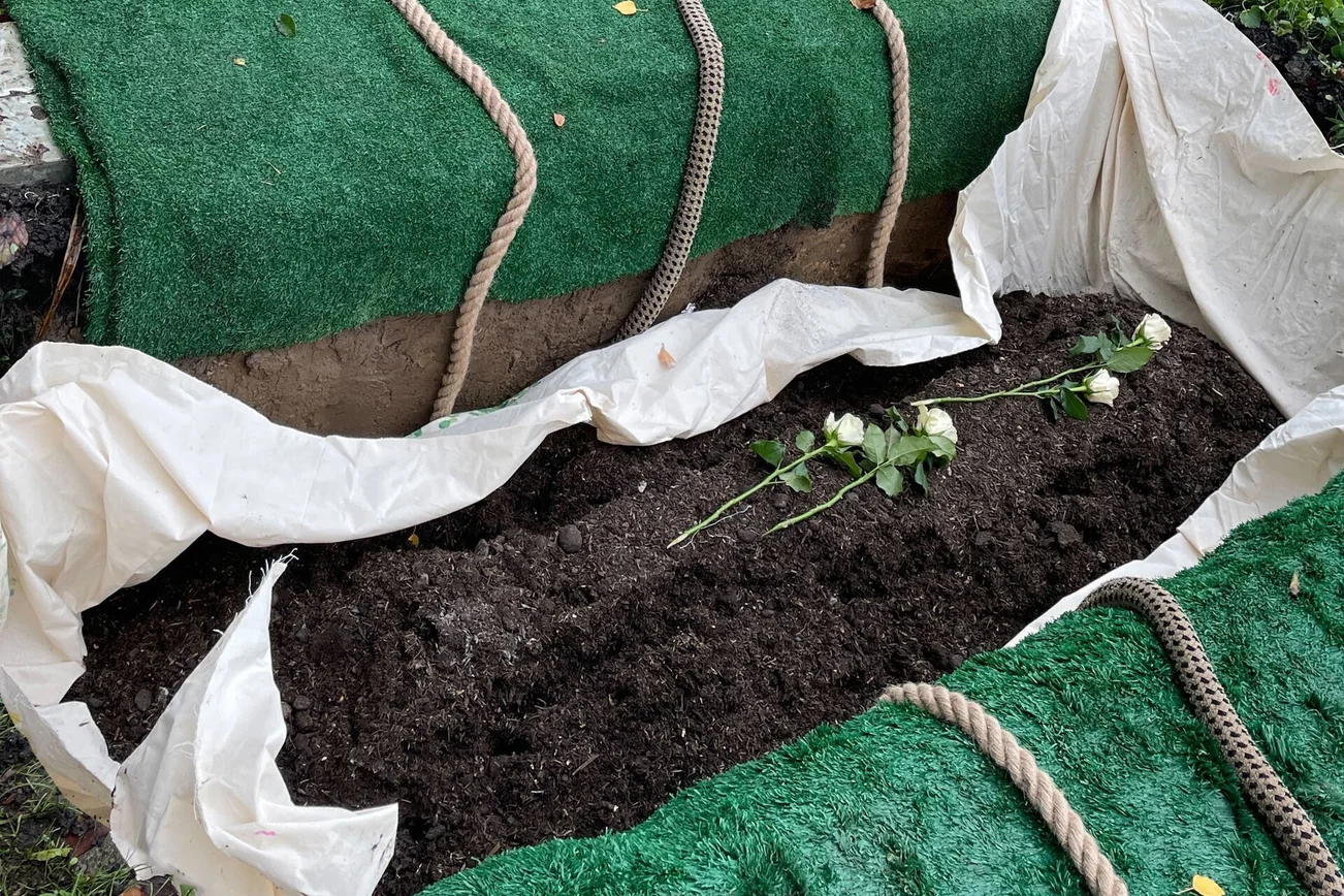 Ein offenes Grab mit einem weißem Tuch und dunkler Erde, darauf mehrere weiße Rosen