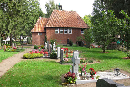 Vor der Friedhofskapelle sind mehrere Gräber, mit blühenden Pflanzen und ein Weg zu sehen - Copyright: Ev.-Luth. Kirchenkreis Lübeck-Lauenburg