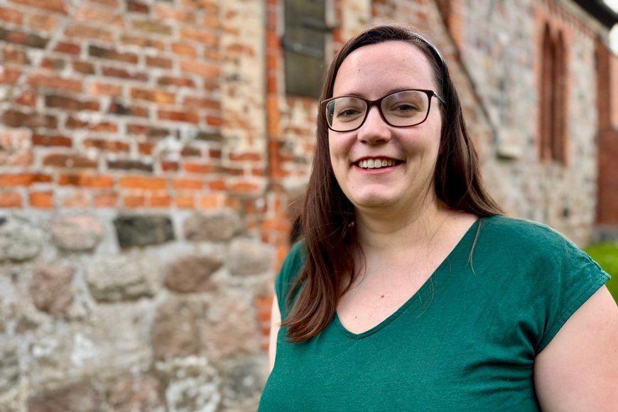 Junge Frau lächelt vor einer Backsteinkirche stehend in die Kamera. - Copyright: Bastian Modrow