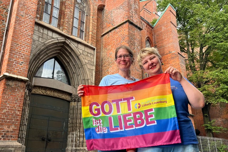 Zwei Frauen stehen vor einer Kirche und halten eine Regenbogen-Fahne mit der Aufschrift 'Gott ist die Liebe' - Copyright: Katharina Micheel