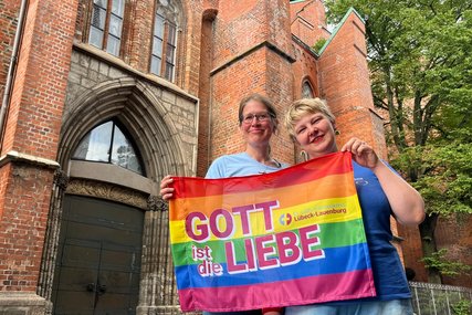 Zwei Frauen stehen vor einer Kirche und halten eine Regenbogen-Fahne mit der Aufschrift 'Gott ist die Liebe' - Copyright: Katharina Micheel
