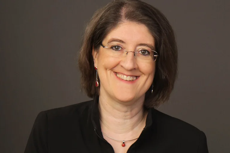 Eine Frau mit braunen mittellangen Haaren schaut lächelnd in die Kamera. Der Hintergrund ist grau-braun verschwommen